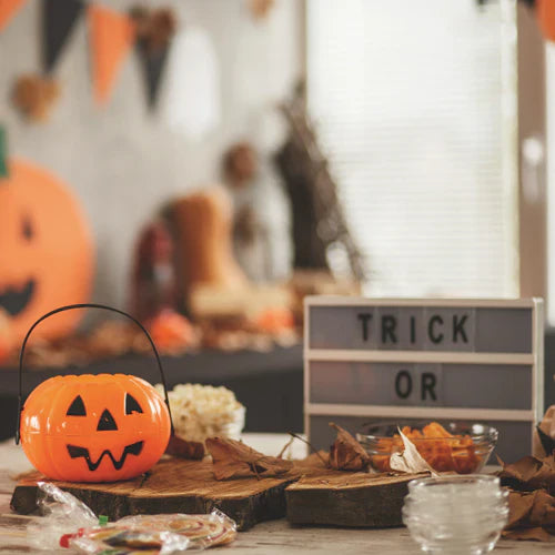 Halloween-themed table with a pumpkin-shaped bucket, candy, autumn leaves, and a "TRICK OR" lightbox sign. Festive decorations are in the background, creating a fun and spooky atmosphere.